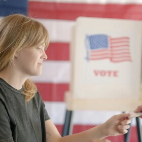 voter worker looks at the ID of a voter in a precinct setting, voting booths in background 