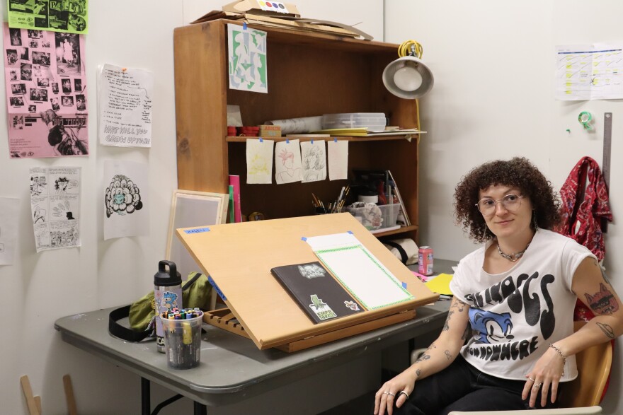 A cartoonist sits at her desk in a studio filled with drawings, posters, markers and sketching supplies.