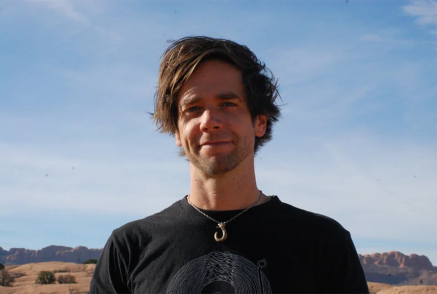 A man with medium-length brown hair stands outside. He wears a black shirt and a hook necklace. He looks at the camera and smiles. 