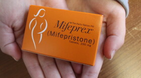 Two hands hold an orange box of pills labeled Mifeprex (Mifepristone) above a wood tabletop 
