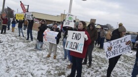 Crowd of protesters standing on snowy ground in front of a store holding various signs that read 'End Violence', 'Abolish ICE', and 'Justice for Renee Good'