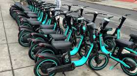 A dozen Veo rental bikes lined up at a rack on a Syracuse sidewalk.