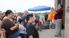 A man stands in front of a crowd holding a weather instrument. He is explaining to the crowd weather balloons work right before he launches one.