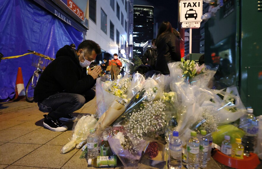 A mourner prays in front of offerings near a building where a fire broke out, in Osaka, western Japan Saturday, Dec. 18, 2021. Japanese police on Sunday identified a 61-year-old man as a prime arson suspect after a fire engulfed a mental clinic in the eight-story building where he was a patient, killing more than 20 people who were trapped inside. (Kyodo News via AP)