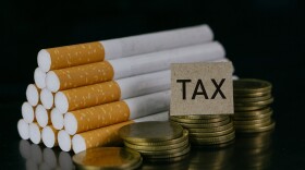 A pile of cigarettes next to growing stacks of coins, with a paper that says "tax"
