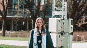 A smiling young woman in graduation regalia stands in front of a large piece of forecasting equipment