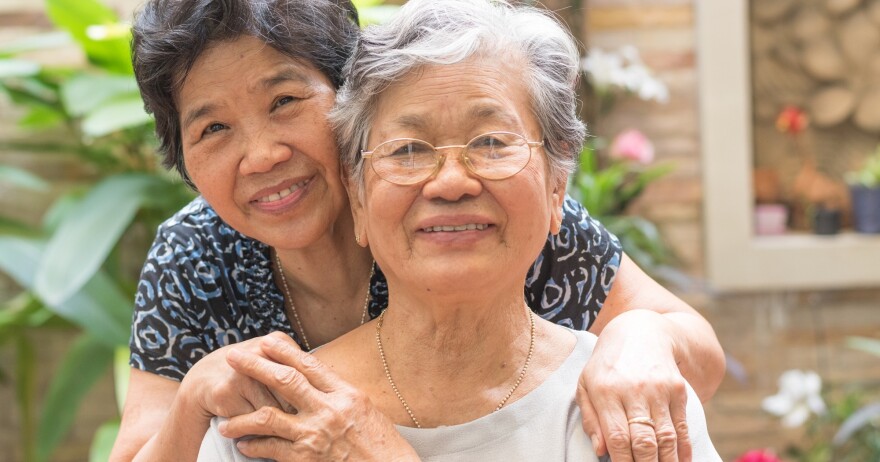 An elderly woman smiles at the camera with her daughter standing behind her.