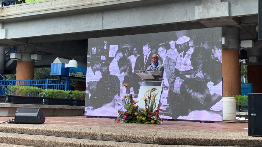 Rodney Hurst speaking on a platform, with a picture of the young black demonstrators in the background, the podium that Hurst is speaking on has flowers in front of it.