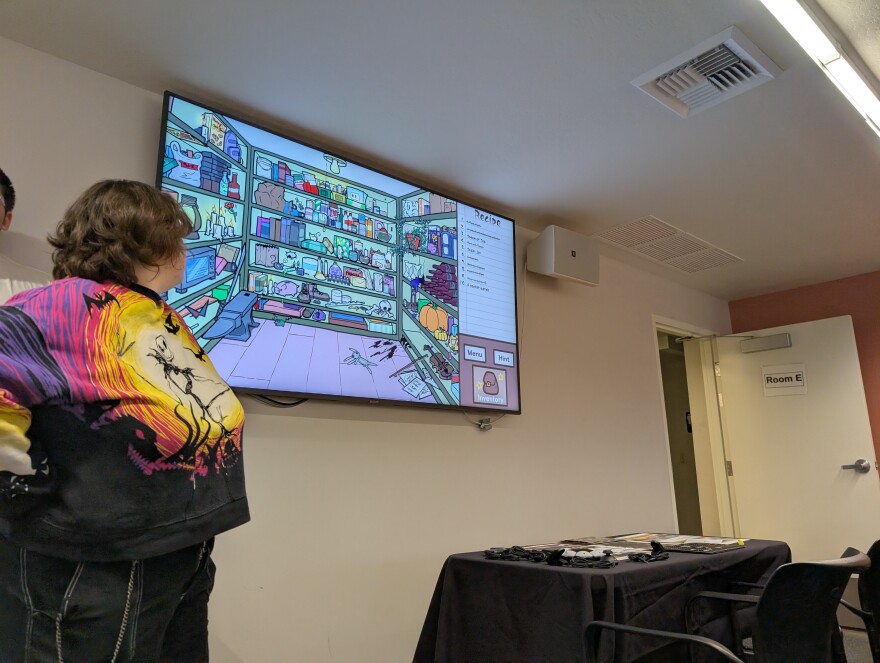 Someone standing on the left looks at a TV screen behind them, with an item collection video game showing
