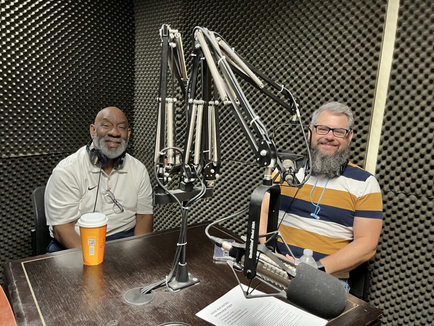 Two men sit smiling at a table in a recording studio.