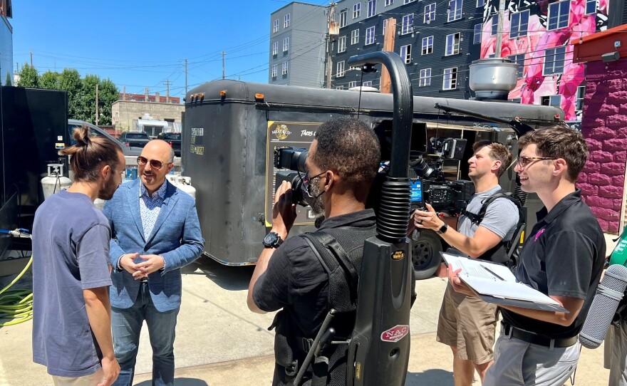 A three-person television crew films a host in sunglasses and a blue blazer interviewing a man with long hair and a beard in a food-truck park.