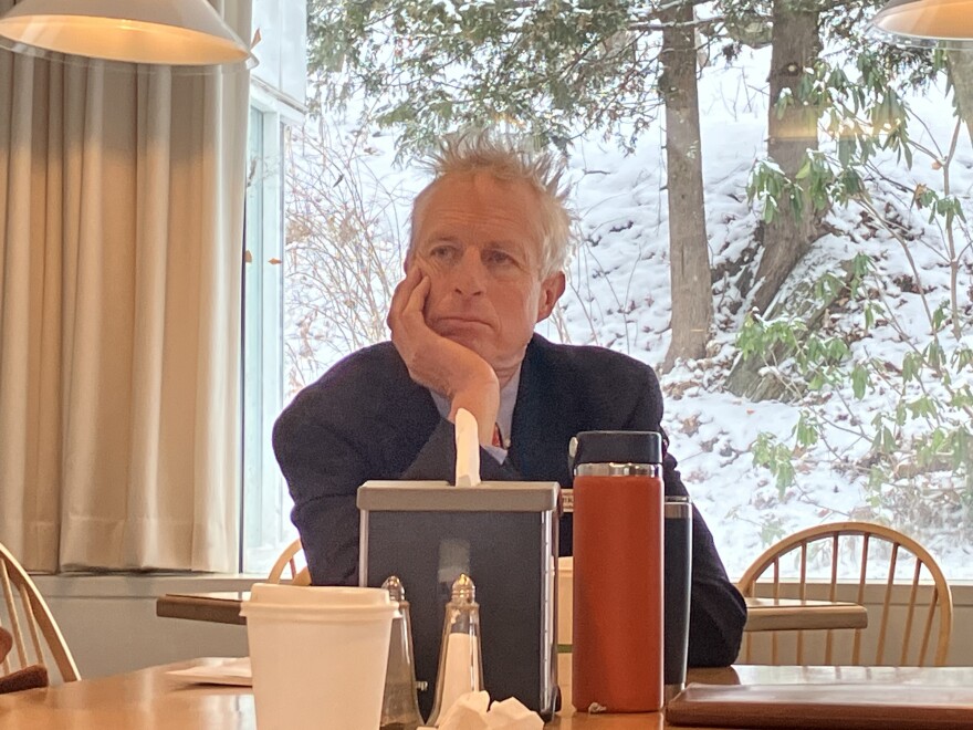 A person in a suit and tie sitting at a table in front of a large window with their hand on their chin