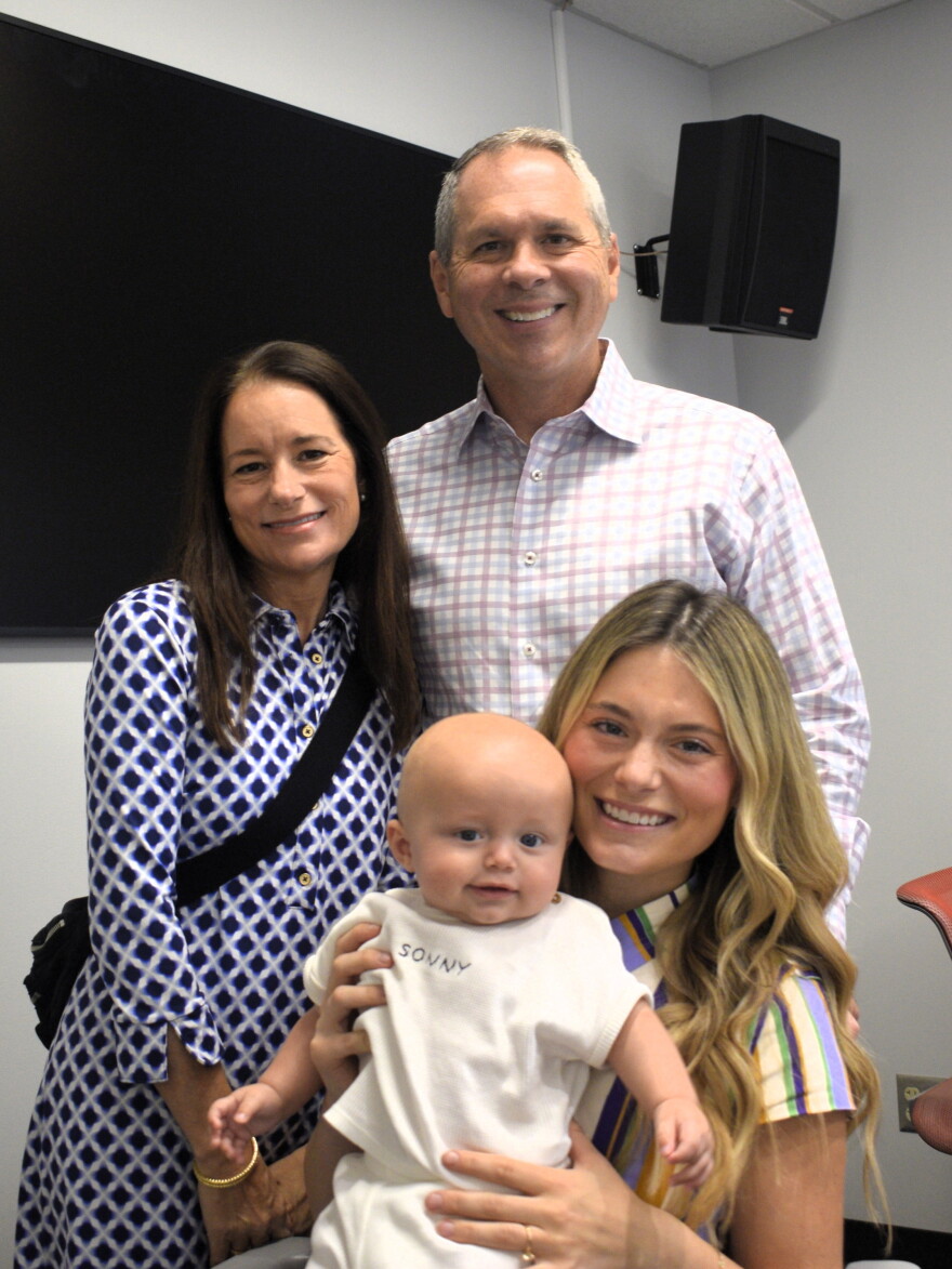Four people pose indoors: a smiling man and woman stand behind a seated woman holding a baby. The baby wears a white outfit, and all appear happy. There’s a black screen and speaker on the wall behind them.