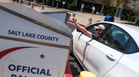 A person leans out of their car in a parking lot to drop a ballot into a drop box, labeled Salt Lake County Official Ballot Drop Box.