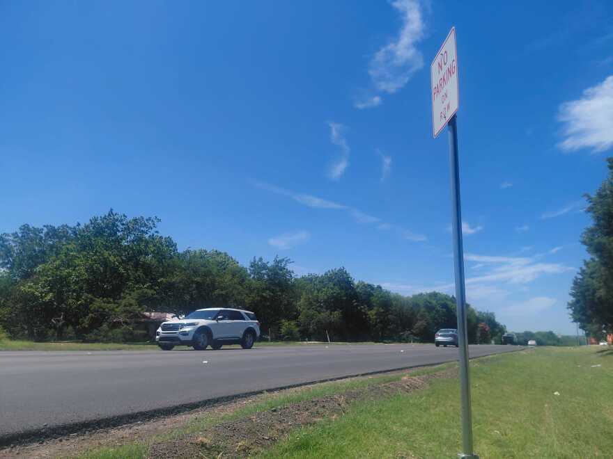 Cars drive in opposite direction on a black asphalt road lined with green grass. A sign in the foreground reads "No parking"