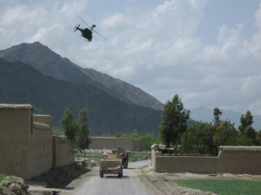 Army vehicles drive down a road in Kunar Province, surrounded by tall mountains. And with a helicopter overhead.