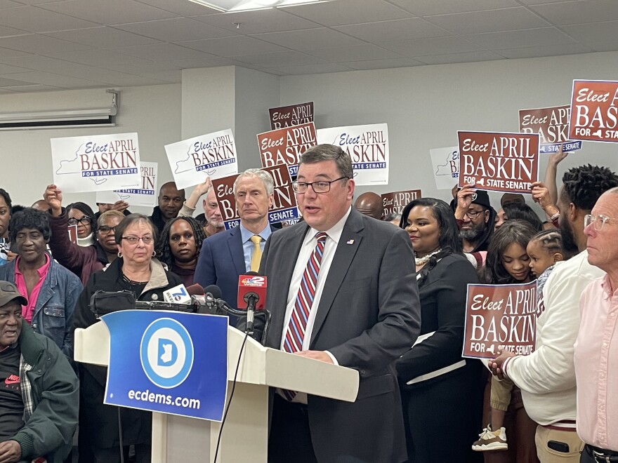 Erie County Democratic Committee Chair and Erie County Board of Elections Commissioner, Jeremy J. Zellner introduces April Baskin as the Democrats' nominee for state senate. Zellner is a white man standing behind a white lectern. He wears a dark grey suit with a white shirt and a red, white and blue striped tie. Behind him stands April Baskin along with her supporters.