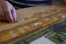 An older hand holds a puzzle piece, next to rows of an in-the-works puzzle with a dirt road, chickens and a farm scene.