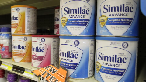 Cans of Similac baby formula sit on a grocery store shelf. The cans are yellow, red and blue. 