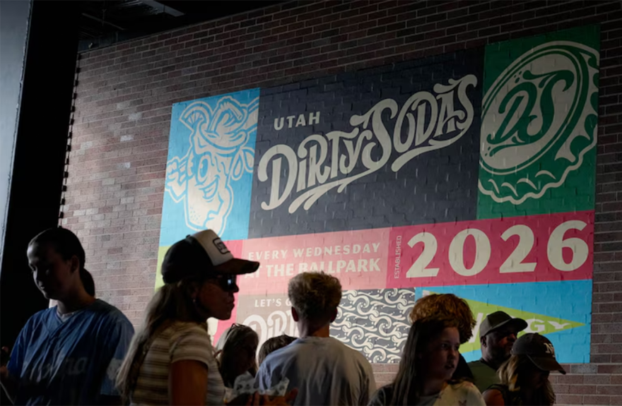 A colorful mural on a brick wall reads "Utah Dirty Sodas" and "2026." A crowd of people stand in front of it, facing different directions. 