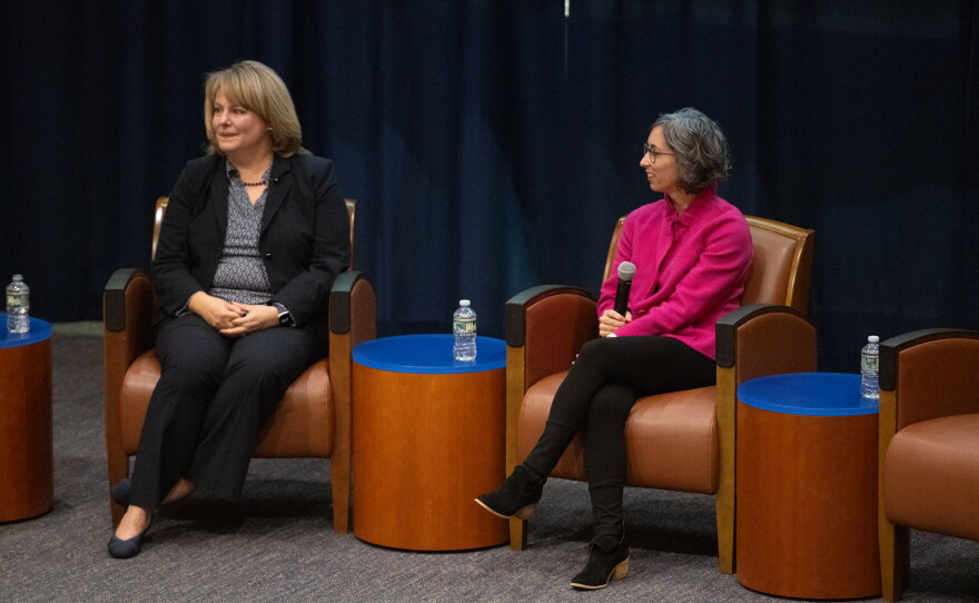 Jenny Steadman sits on a chair next to Michelle Riordan-Nold. Michelle is holding a microphone and they are looking off to the side as they participate in a live panel discussion.