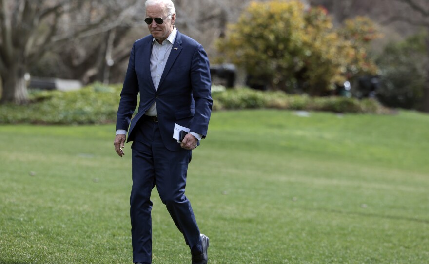President Biden walks across the lawn, holding his phone and paper in one hand.