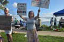 A woman wearing a white shirt and muted yellow floral skirt is holding a sign that says "Trump for Prison 2025" in her left hand and other sign in her right. There are other people and signs to her left and back.