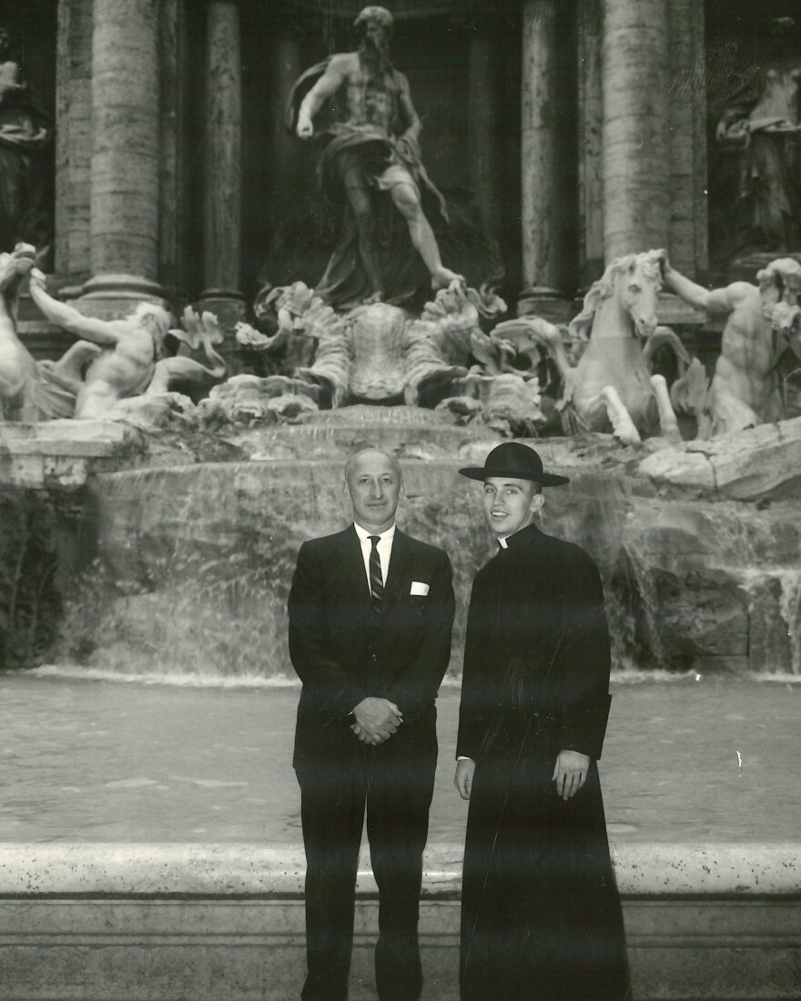 Two men stand in front of the Trevi Fountain in Rome; one wears a suit and tie, the other is dressed in a priest’s cassock and hat. The large fountain and sculpted figures are visible behind them.