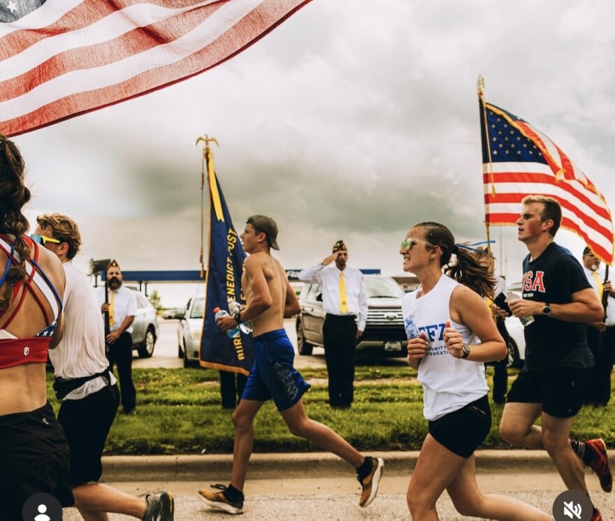 Runners participate in a race, some holding American flags. People in dress shirts stand nearby, and cloudy skies are overhead. The scene appears patriotic and energetic, with flags waving prominently in the background.