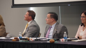 At a black table, Russ Kavalhuna sits at an angle, smiling at someone off camera. He is wearing a grey suit with a yellow tie. Next to him, Alan Turfe also looks off camera. He is wearing a grey suit with a purple tie. Next to them, board secretary Stacey Thinnes is pictured in a light pink suit with arms crossed. 