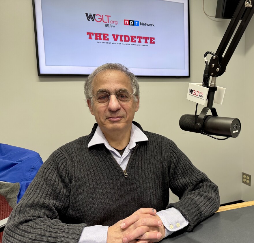 Person sitting at a desk in a radio studio, wearing a black sweater and glasses, with a microphone in front. A sign on the wall behind reads "THE VIDETTE" and includes logos for WGLT and NPR Network.