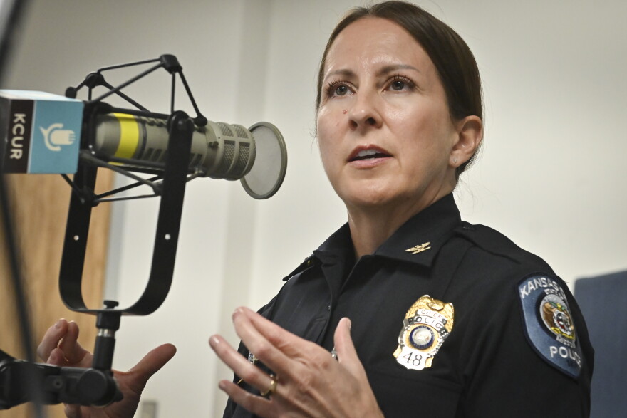 A woman wearing a Kansas City Police uniform sits inside a radio studio. She gesturing with both hands and talking at a microphone.