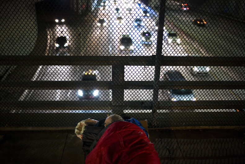 A homeless man lies tucked in a sleeping bag on an overpass above the Freeway.