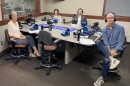 Four smiling people wearing headphones sit around a table in a radio talk studio: a woman front left has short grey hair and is wearing glasses, a white blouse, dark pants, and tan shoes; a woman back left has long light brown hair and is wearing a grey sweater; a man back center has short dark hair and a dark mustache and beard and is wearing a white button-down shirt under a grey suit coat; a man at right has very short dark hair and is wearing glasses, a blue button-down shirt, jeans, and grey and blue sneakers