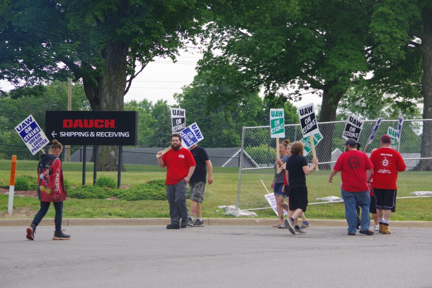 Around ten people stand on the side of the road in front of a chainlink fence and a black sign to the middle right of the image that reads "DAUCH" in red text, below which reads "Shipping & Receiving" in white text with an arrow pointing to the right. The men and women hold different colored signs, but most of them read the same message: "UAW ON STRIKE." One woman's sign to the left of the image reads "UAW ON STRIKE, UNFAIR LABOR PRACTICE" in blue text.