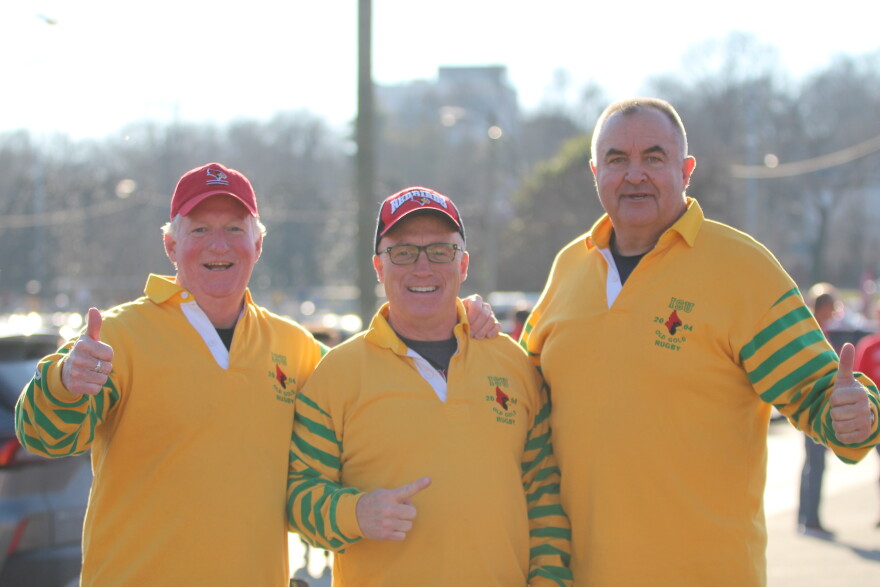 Three smiling men wearing yellow rugby shirts with green-striped sleeves and red caps stand together outdoors, giving thumbs up in a sunny setting. Trees and cars are visible in the blurred background.