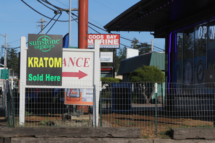 A strip mall during the day. A sign reads "Kratom Sold Here" outside of a smoke shop in a strip mall.
