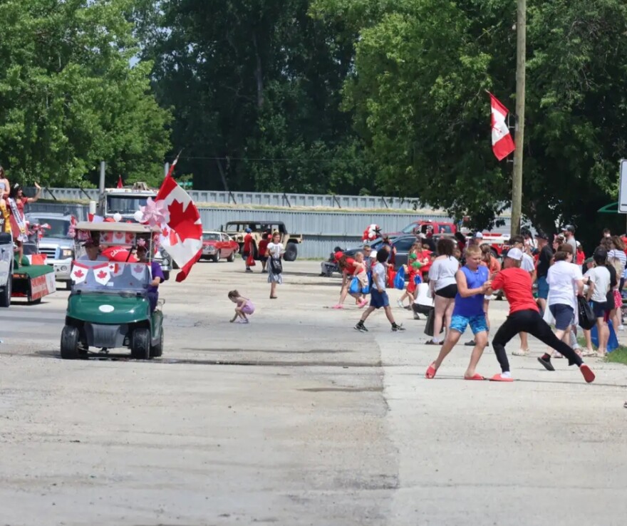 People line up on the sidewalk as a parade of cars, carts and floats drive down Main Street in Emerson, Manitoba | MPR News