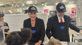 Michigan Governor Gretchen Whitmer and state Senator Sean McCann serve school meals to children at a cafeteria, wearing matching “Champion of the Tray” caps and “Nutrition’s Game One” aprons.
