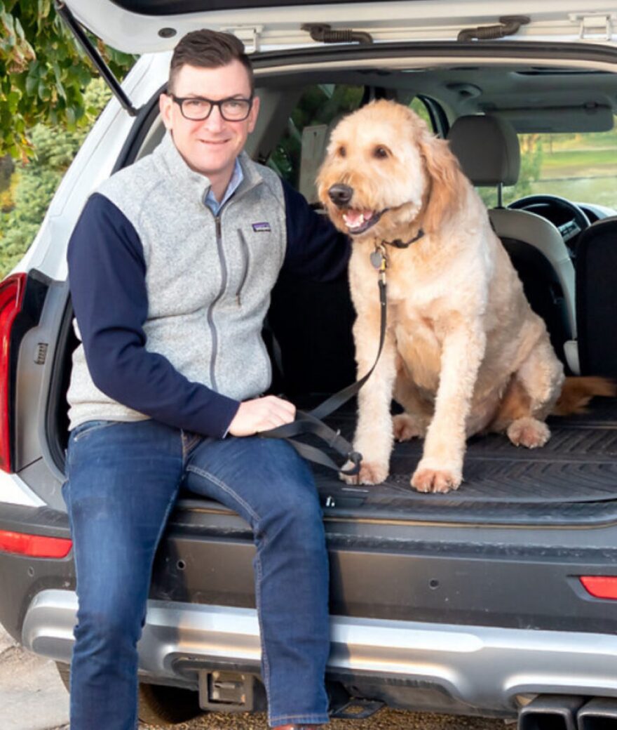 Man with brown hair and glasses in car with big golden-doodle looking dog 