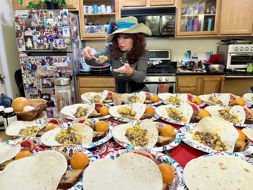 A table is covered with paper plates filled with food. An older woman with long brown hair and a large hat spoons food onto a plate. 