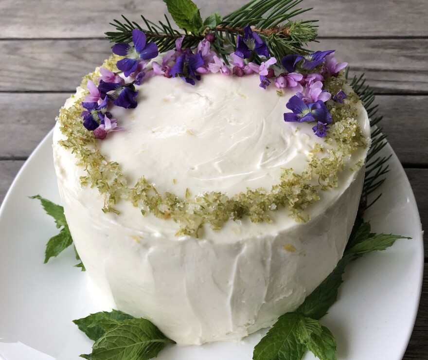 white layer cake with purple flowers, spruce sprigs and green sugar decoration plus mint leaves