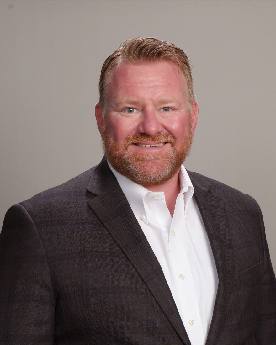 A smiling man with short red hair and a red beard wearing a black blazer and a white button-down shirt.