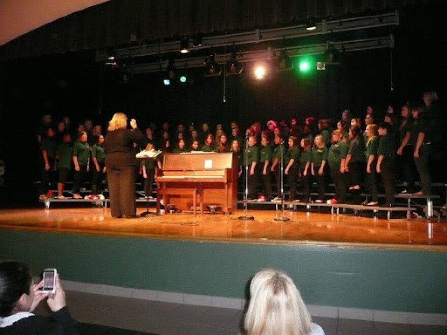 A woman with blonde hair seen from behind, conducting singers