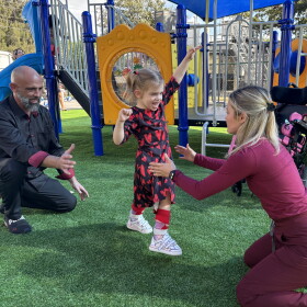 A young toddler walks from one adult toward another on a playground