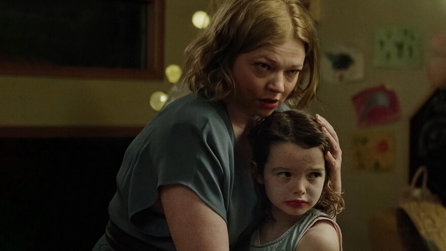A still image from a movie shows a woman with a concerned look on her face holding her young daughter close as they stand in the family room of a home.