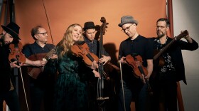 Six musicians pose together in a studio, holding string instruments.