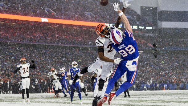 A football player in an orange helmet jumps to block another football player in a blue uniform from catching a ball. 
