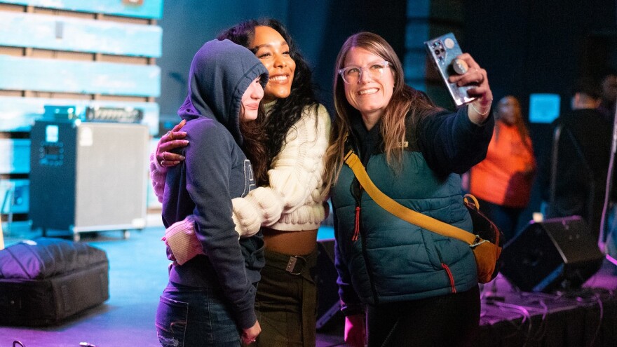 Three women pose for a selfie, one holding a phone with her arm extended and the other two embracing.