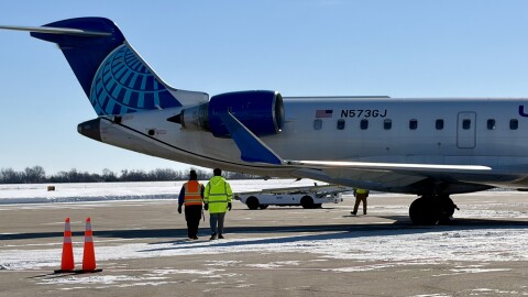 Two men dressed in neon yellow and orange walk towards the back of a United Airlines aircraft parked at the gate. 